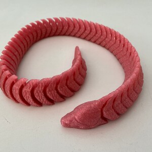 Può includere: Un braccialetto di plastica rosa flessibile e segmentato con una superficie testurizzata. Il braccialetto ha la forma di un serpente ed è arrotolato in un cerchio.