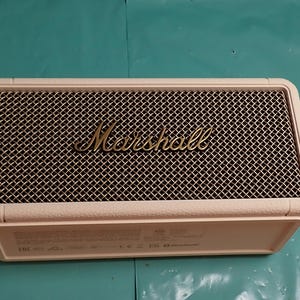May include: A cream-colored Marshall speaker with a black mesh front and gold lettering. The speaker has a rectangular shape with rounded edges. The Marshall logo is prominently displayed on the front. The speaker is designed for audio playback.