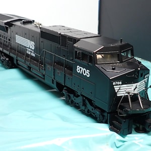 May include: A black model train locomotive with the number 8705 on the front and side. The train has the words "NS SOUTHERN" printed on the side in white. The train is on a light blue surface.