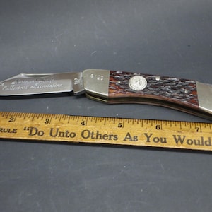 May include: A folding pocket knife with a stainless steel blade and a brown handle. The blade is engraved with text. A wooden ruler with text is in the foreground.