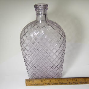 May include: A clear, vintage-style glass bottle with a diamond pattern. The bottle has a rounded body and a long neck. The bottle is approximately 9 inches tall. The bottle is a light purple color.
