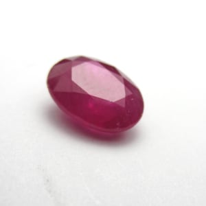 May include: A faceted, oval-shaped ruby gemstone. The gem is a deep, saturated red color, with multiple facets that catch the light. The ruby is set against a white background.