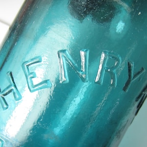 May include: Close-up of a teal-colored glass bottle with the embossed word "HENRY". The bottle has a textured surface and is partially visible, with the text appearing in a slightly lighter shade of teal.