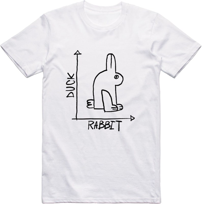 May include: White t-shirt with a black line drawing of a rabbit that could also be a duck. The words "DUCK" and "RABBIT" are written on the shirt, along with an arrow.