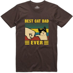May include: Brown t-shirt with a retro-style graphic that says "Best Cat Dad Ever". The graphic features a fist bumping a cat with a black and white face.