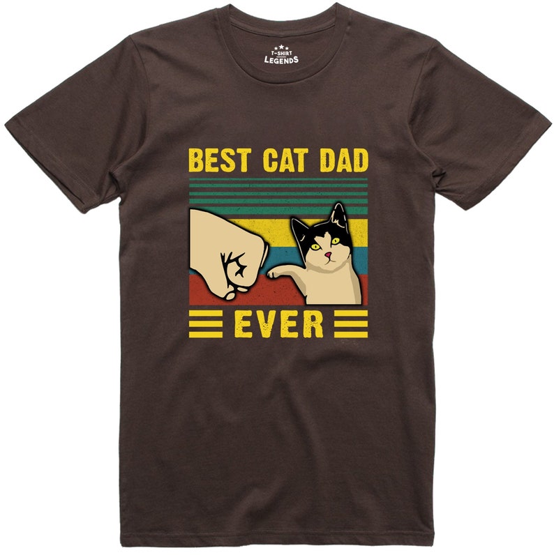 May include: Brown t-shirt with a retro-style graphic that says "Best Cat Dad Ever". The graphic features a fist bumping a cat with a black and white face.