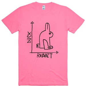 May include: A bright pink t-shirt with a black graphic of a rabbit, with the words "DUCK" and "RABBIT" in black text. The rabbit is drawn in a minimalist style, with a single eye and simple lines. The shirt is made of a soft material.