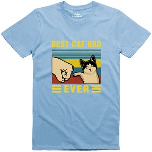 May include: Light blue t-shirt with a retro-style graphic of a cat pawing at a fist. The text "Best Cat Dad Ever" is printed in yellow on the shirt.