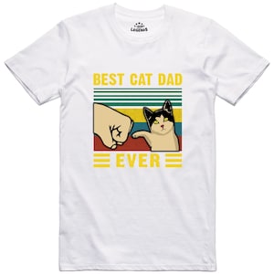 May include: White t-shirt with a retro-style graphic that says "Best Cat Dad Ever". The graphic features a fist bumping a cat with a black and white face.