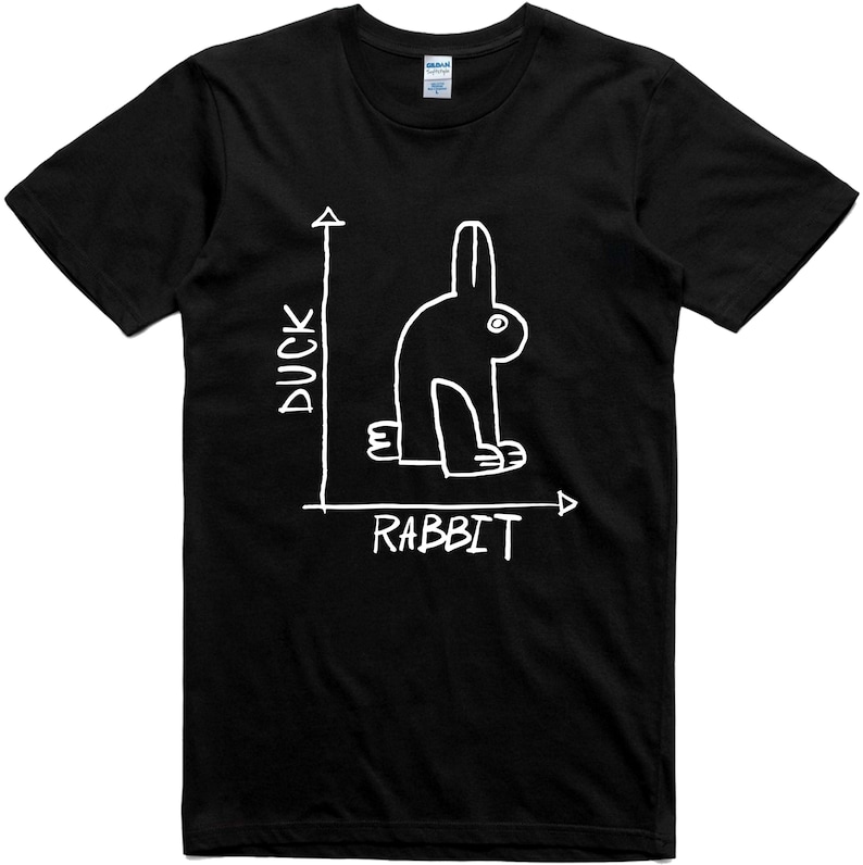 May include: Black t-shirt featuring a white line drawing of a rabbit, with the words "DUCK" and "RABBIT" in white. The design is a play on the classic duck-rabbit optical illusion. The shirt is made of a soft material.