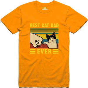 May include: A yellow t-shirt with a retro-style graphic that says "Best Cat Dad Ever". The graphic features a cat giving a fist bump to a hand.