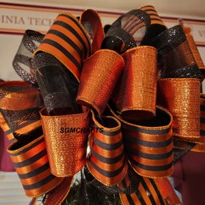 May include: A decorative bow featuring multiple loops of ribbon in metallic orange, solid black, and orange-and-black striped patterns. The ribbon has a shimmering texture and includes a black mesh material. The text SGMCRAFTS is visible on one loop.