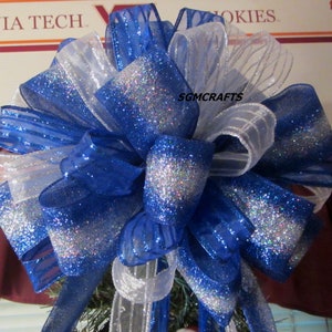 May include: A blue and white glittery Christmas tree topper bow. The bow is made of two types of ribbon, one with silver glitter and one with white stripes. The bow is tied with a white ribbon.