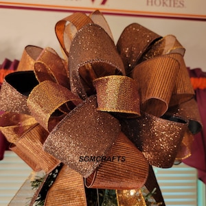 May include: A large, decorative bow made of various brown ribbons. Some ribbons have a glittery finish, while others are textured. The bow is likely for holiday or gift decoration. The text "SGMCRAFTS" is visible on the bow.