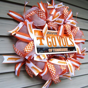 May include: A decorative wreath featuring orange and white ribbons, mesh, and a license plate that reads "GO VOLS" and "UNIVERSITY OF TENNESSEE". The wreath has a checkerboard pattern and is designed for door or wall decor.