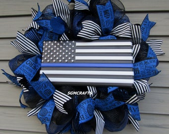 May include: A blue and black wreath with a thin blue line American flag in the center. The wreath is decorated with ribbons that have the words "Protect & Serve", "Hero", "Peace", "Brave", "Blue", "Enforcer", "Police", "Strong", and "2007" printed on them.