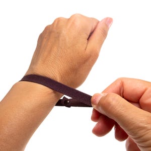 Könnte beinhalten: Ein dunkelbraunes elastisches Armband mit einem kleinen, runden Metalldruckknopf. Das dünne, flexible Stoffband ist so konzipiert, dass es um das Handgelenk gelegt werden kann, wobei der Druckknopf als sicherer Verschluss dient.