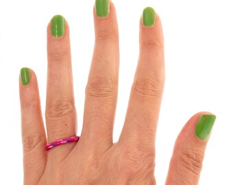 May include: A thin, circular ring featuring a vibrant pink and purple braided cord texture. The band is constructed from a flexible, woven material with a consistent cylindrical shape, worn on the ring finger of a hand with bright green nail polish.