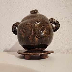 May include: A round, dark brown glazed ceramic vessel with a narrow neck and two side handles featuring circular cutouts. The vessel rests on a matching circular, three-legged ceramic base, showcasing a glossy, mottled finish throughout.