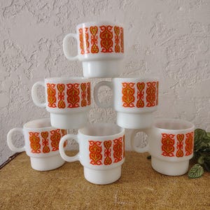 May include: A set of five white milk glass mugs stacked in a pyramid formation. Each mug features a repeating orange and yellow decorative pattern around the exterior. The mugs have a cylindrical shape with a stepped base and a side handle.