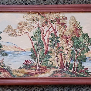 May include: A framed landscape painting featuring a serene lake scene with trees in shades of green, red, and brown. The artwork is enclosed in a reddish-brown wooden frame. The painting depicts a path leading to the lake.