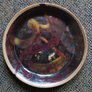 May include: A round ceramic bowl with a dark brown interior and a light tan rim. The bowl's interior features abstract yellow and maroon designs. The bowl is set against a textured gray surface.