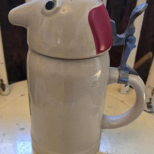 Peut inclure: Une chope à bière en céramique beige avec un couvercle à charnière. Le couvercle est en forme de tête d'oiseau avec un accent rouge. La chope a une poignée en métal et un marquage de capacité de 0,5L. La chope est posée sur une surface blanche.