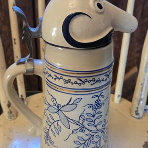 Peut inclure: Une chope à bière en céramique vintage au design fantaisiste. La chope blanc cassé est dotée d'un couvercle avec un dessus en métal argenté et d'une poignée. Le corps est décoré de motifs floraux bleus et de bandes bleues et dorées.