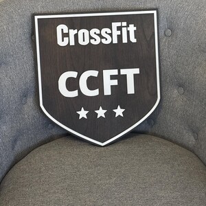 May include: A dark brown wooden shield-shaped sign with white text that reads "CrossFit CCFT" and three white stars.
