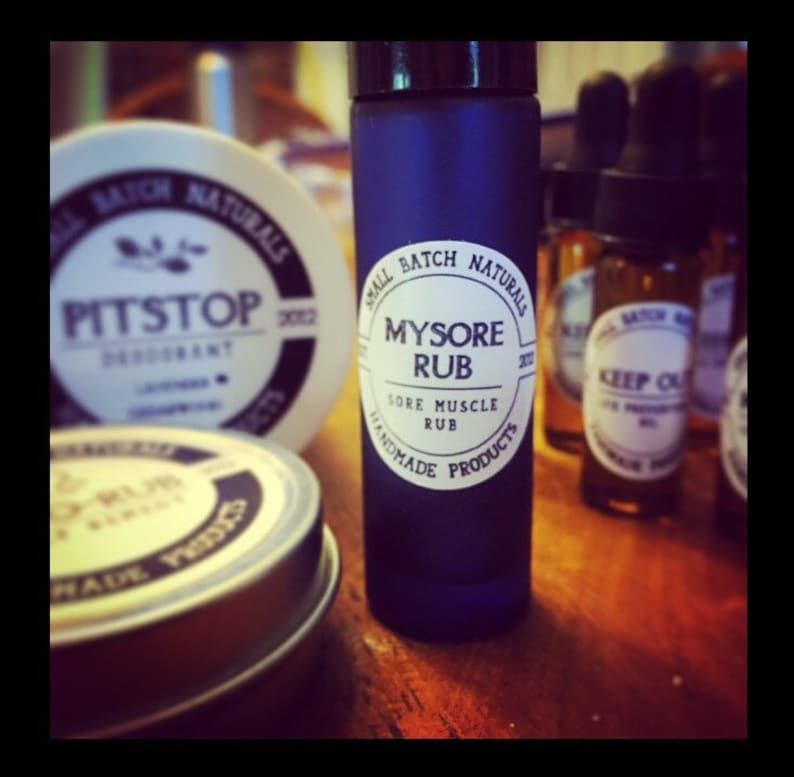 Puede incluir: Una botella de vidrio azul con una etiqueta blanca que dice "Small Batch Naturals Mysore Rub Sore Muscle Rub Handmade Products 2012". La botella est&aacute; sentada sobre una superficie de madera con otras botellas.