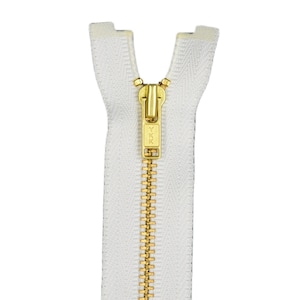 Puede incluir: Una cremallera blanca con un deslizador y una lengüeta de metal dorado. La cremallera está cerrada y el deslizador tiene el texto "YKK" en él.