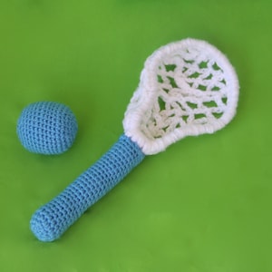 May include: A blue and white crocheted lacrosse stick and ball. The stick has a white net and a blue handle. The ball is blue.