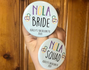 May include: Two round blue and white buttons with the text "NOLA Bride" and "NOLA Squad" with purple and yellow accents. The text "Ashley's Bachelorette 2020" is printed below the text on each button.