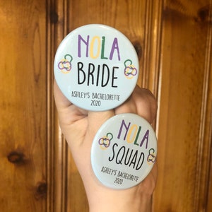 Peut inclure: Deux boutons ronds bleus et blancs avec le texte "NOLA Bride" et "NOLA Squad" avec des accents violets et jaunes. Le texte "Ashley's Bachelorette 2020" est imprimé sous le texte de chaque bouton.
