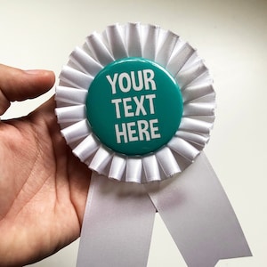May include: A white rosette with a teal center button. The button has the text "YOUR TEXT HERE" in white. The rosette has white pleated fabric around the button and two white ribbon tails.