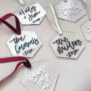 May include: Six clear acrylic hexagon ornaments with black and white calligraphy. The ornaments are decorated with names and dates, including "Holly & Jim 11-1-20", "the Cuomos 2020", "the Millers 2020", "Buckley 2020", "baby Toole 2021".
