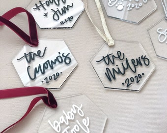 May include: Six clear acrylic hexagon ornaments with black and white calligraphy. The ornaments are decorated with names and dates, including "Holly & Jim 11-1-20", "the Cuomos 2020", "the Millers 2020", "Buckley 2020", "baby Toole 2021".