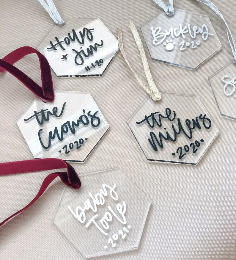 May include: Six clear acrylic hexagon ornaments with black and white calligraphy. The ornaments are decorated with names and dates, including "Holly & Jim 11-1-20", "the Cuomos 2020", "the Millers 2020", "Buckley 2020", "baby Toole 2021".