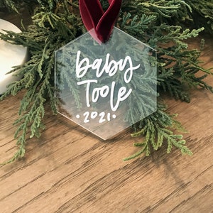 May include: A clear hexagonal ornament with a red ribbon. The ornament has white lettering that says "Baby Toole - 2021".