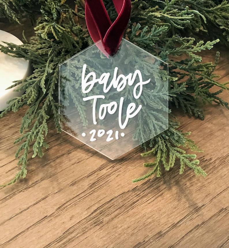 May include: A clear hexagonal ornament with a red ribbon. The ornament has white lettering that says "Baby Toole - 2021".