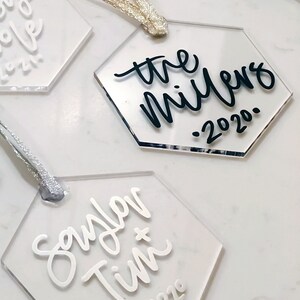 May include: Three clear acrylic hexagon ornaments with white and black calligraphy. The first ornament says "the millers . 2020.", the second ornament says "saylor + tim 2020", and the third ornament says "our love 2021".