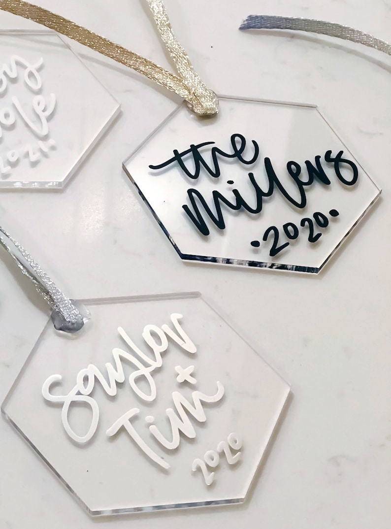 May include: Three clear acrylic hexagon ornaments with white and black calligraphy. The first ornament says "the millers . 2020.", the second ornament says "saylor + tim 2020", and the third ornament says "our love 2021".