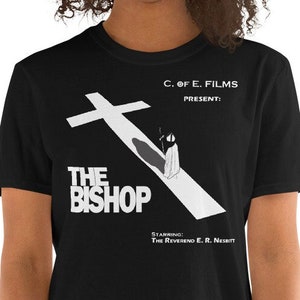 May include: Black t-shirt featuring a white graphic design of a person walking away from a cross. The graphic text reads "C. of E. Films Present: The Bishop Starring The Reverend E.R. Nesbitt"