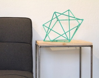 May include: A teal geometric metal sculpture sits on a wooden top side table with a black metal frame. The table is next to a dark gray couch.