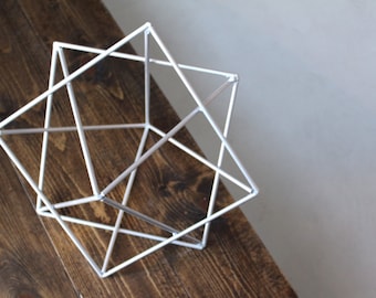 May include: A white geometric sculpture made of thin metal rods, resembling a cube with intersecting lines. The sculpture is resting on a wooden surface.