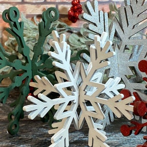 May include: A set of three white wooden snowflakes with a glittery finish. The snowflakes are layered on top of each other and are arranged in a starburst pattern.