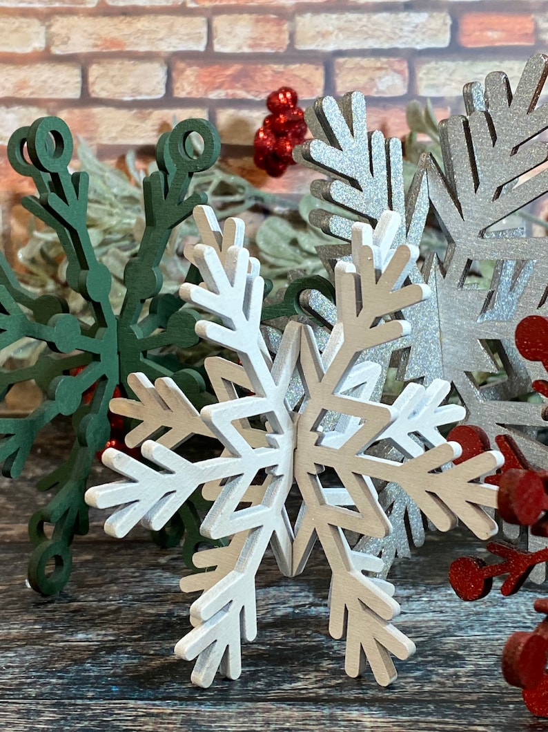 May include: A set of three white wooden snowflakes with a glittery finish. The snowflakes are layered on top of each other and are arranged in a starburst pattern.
