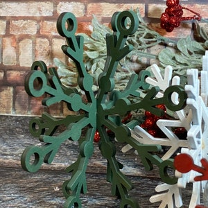 May include: A dark green wooden snowflake ornament with a 3D design. The snowflake has a circular hole in the center and is made of multiple layers of wood.