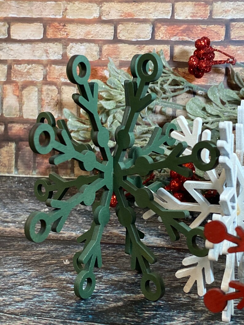 May include: A dark green wooden snowflake ornament with a 3D design. The snowflake has a circular hole in the center and is made of multiple layers of wood.