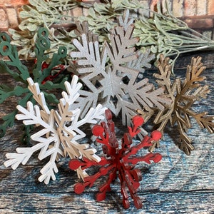 May include: Six wooden snowflake ornaments in various colors, including green, silver, white, gold, and red. The snowflakes are arranged on a rustic wooden surface.